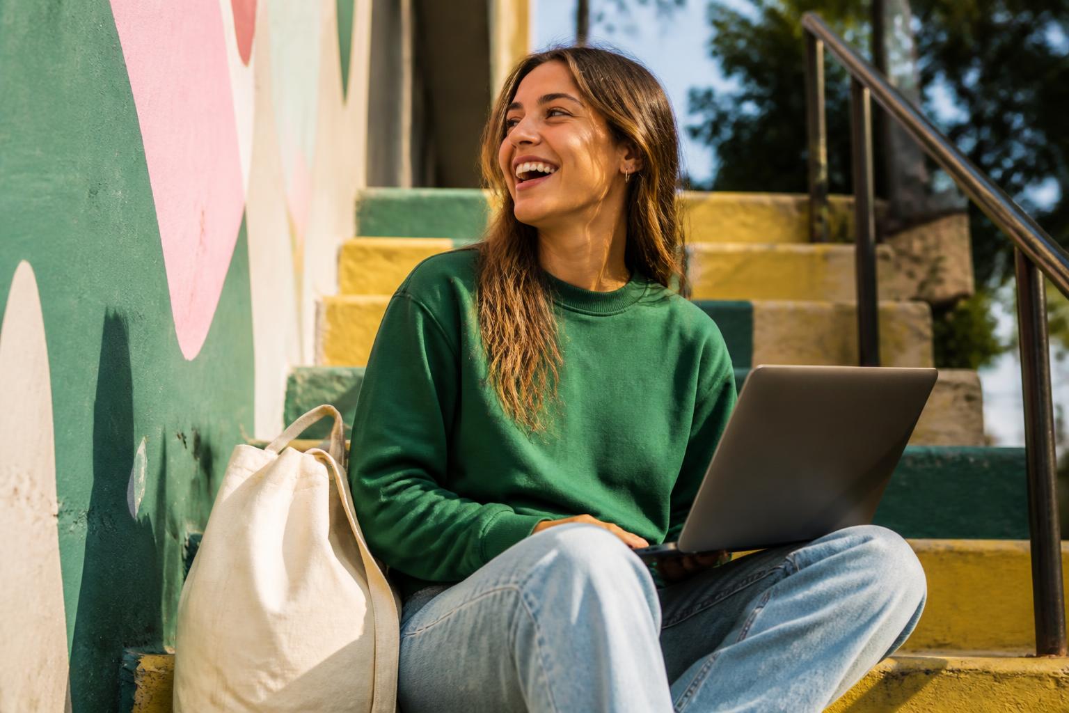 A recent graduate laughing on colourful steps with a laptop